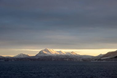 Norveç denizinin sakin suları. Karla kaplı dağlar, kutup gündoğumunda parlak sarı renkler. Renkli gökyüzü. Harstad, Norveç.