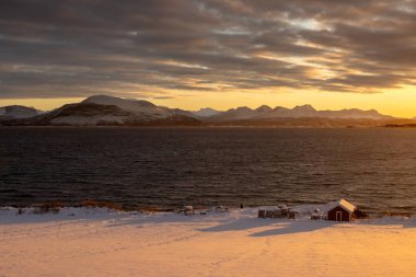 Kuzey Kutbu sonbaharında soğuk bir günde gün doğumu. Doğa karla kaplı. Ufuktaki dağlar. Sahildeki kırmızı kulübe. Bulutlu renkli gökyüzü. Lundenes, Grytoya Adası, Norveç.