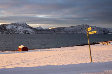 Kuzey Kutbu sonbaharında soğuk bir günde gün doğumu. Doğa karla kaplı. Ufuktaki dağlar. Sahildeki kırmızı kulübe. Bulutlu renkli gökyüzü. Lundenes, Grytoya Adası, Norveç.