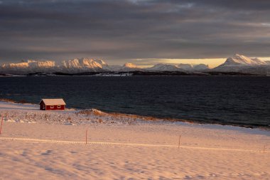 Kuzey Kutbu sonbaharında soğuk bir günde gün doğumu. Doğa karla kaplı. Ufuktaki dağlar. Sahildeki kırmızı kulübe. Bulutlu renkli gökyüzü. Lundenes, Grytoya Adası, Norveç.