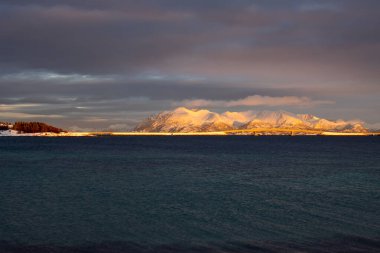 Norveç denizinin sakin karanlık suları. Ufukta karlı dağlar. Güneşin doğuşunda renkli bulutlu bir gökyüzü. Lundenes, Grytoya, Norveç.