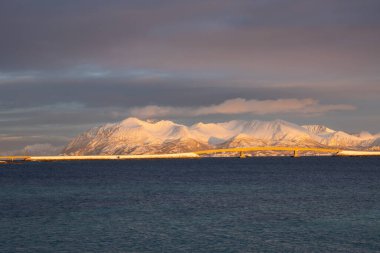 Norveç denizinin sakin suları. Ufukta kar görünen dağlar güneş ışığıyla aydınlanır. Güneşin doğuşunda renkli bulutlu bir gökyüzü. Tjeldevika, Grytoya, Norveç.