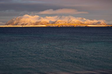 Norveç denizinin sakin suları. Ufukta kar görünen dağlar güneş ışığıyla aydınlanır. Güneşin doğuşunda renkli bulutlu bir gökyüzü. Tjeldevika, Grytoya, Norveç.