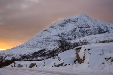 Kırsal yol, kara ve dağ karla kaplı. Kutup güneşinin doğuşunda renkli bir gökyüzü. Rüzgarın kenarında kar ilerliyor. Tjeldevika, Norveç.