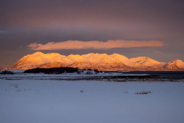 Karla kaplı topraklar. Norveç denizinin suyu. Ufukta karlı dağlar, güneş ışığıyla turuncu renkte. Renkli bulutlu gökyüzü. Lundenes, Harstad, Norveç.
