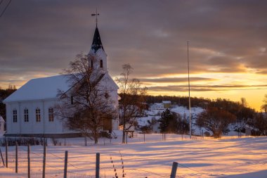Yüksek kulesi olan beyaz ahşap kilise. Kutup günbatımında güneş ışığı, gökyüzünü boyuyor. Karlı arazi. Sandsoya Adası, Norveç.