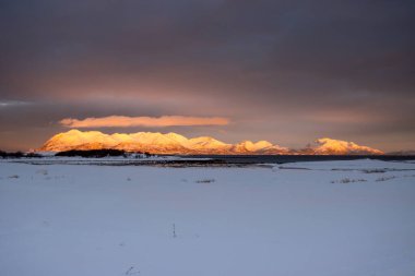 Karla kaplı topraklar. Norveç denizinin suyu. Ufukta karlı dağlar, güneş ışığıyla turuncu renkte. Renkli bulutlu gökyüzü. Lundenes, Harstad, Norveç.