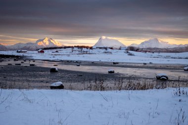 Kuzey Kutbu sonbaharında karla kaplı bir ülke. Büyük kayalarla Norveç denizinin sığ suları. Arka planda dağlar. Gün batımında renkli bir gökyüzü. Lundenes, Grytoya, Norveç.