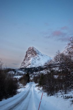 Karla kaplı topraklar. Ufukta karlı Dağ Zirvesi. Yol buzla kaplı. Mavi saatte gökyüzü bulutlu. Lundenes, Grytoya, Norveç.
