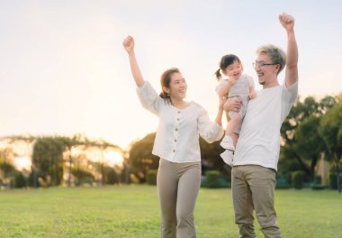 Asyalı anne ve baba kızlarıyla parkta yürürken ebeveynler bebeğinin elini tutup parkın akşam ışığında mutlu bir aile olarak ortaya çıkıyorlar. Mutlu aile kavramı.
