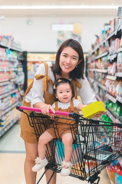 Asian mother and her daughter buying food at huge supermarket ,Hand holding a product box mockup ready for retouch, Baby sit in trolley, Family shopping concept.