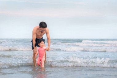 Asyalı baba destek olmak ve sahilde yürümek için el ele tutuşuyor, bebek kız yürümeyi öğreniyor, mutlu aile aktiviteleri konsepti..