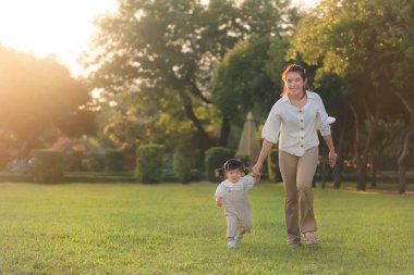 Asyalı bir anne kızıyla parkta yürüyor, ebeveynler bebeğin elini tutuyor, mutlu bir aile parkın akşam ışığında. Mutlu aile kavramı.