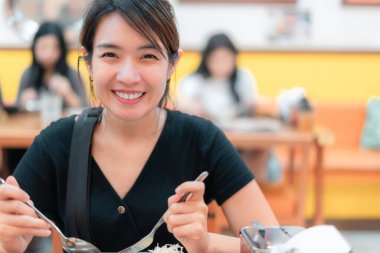 Bir kadın restoranda yemek yerken gülümsüyor. Elinde çatal ve kaşık tutuyor.