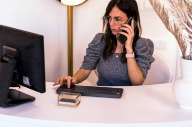 Gözlüklü şık bir kadının cep telefonuyla konuşurken ve güzellik salonunun resepsiyonunda çalışırken bilgisayara veri girerken elde çekilmiş fotoğrafı.