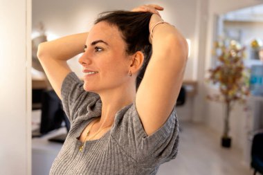 Genç, kendine güvenen, uzun saçlarını elleriyle tutarken gülümseyen esmer kadın.