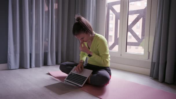 在网上瑜伽课之前 身穿运动服 留着一头长长的黑头发的年轻女性坐在Lotus的垫子上 浏览笔记本电脑 — 图库视频影像