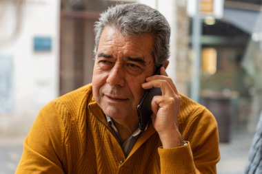 Orta yaşlı, gri saçlı bir adam şehir sokağının bulanık arka planında telefona cevap veriyor.