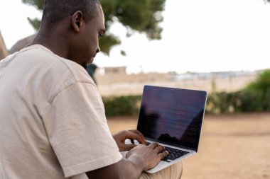 Black man typing on laptop keyboard and creating code while working on freelance project on beach in summer