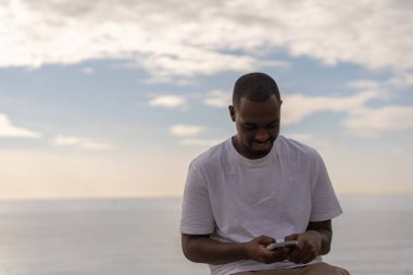 Happy young African American male in casual clothes browsing mobile phone near sea against cloudy sky during sunset