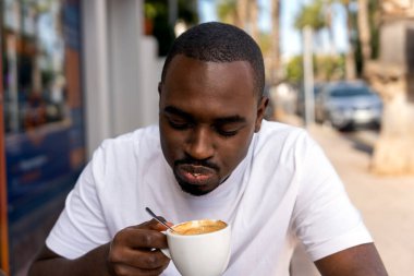 Concentrated young bearded African American male in white t shirt drinking hot coffee in outdoor cafe near palm trees