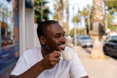 Smiling young bearded African American man in casual clothes sitting at table in outdoor cafe and drinking hot coffee while enjoying cityscape