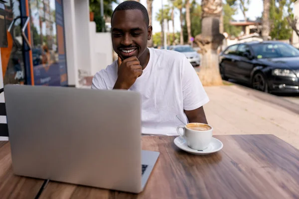 Positive black man in white t shirt touching chin and smiling while sitting at table with cup of latte and analyzing data on laptop