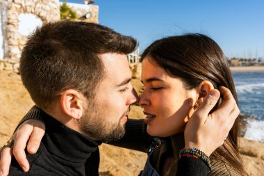 Young man touching hair and kissing girlfriend during romantic shore of waving sea on Saint Valentine Day