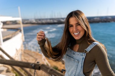 Ethnic lady looking at camera and raising arm while having fun with long hair on pier against sea in sunlight