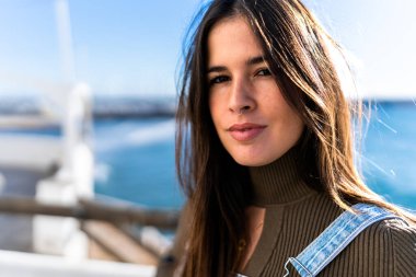 Attractive long haired Hispanic lady looking at camera on blurred background of pier near sea in sunshine