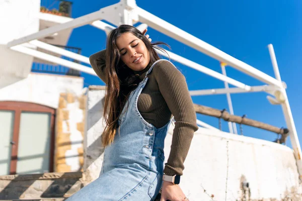 Low angle content ethnic woman in denim overall touching hair with closed eyes while sitting near old pier in sunlight