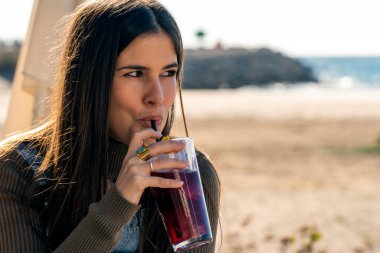 Young female in casual clothes drinking fresh red cold drink through straw from glass while spending time on seashore in sunny weather