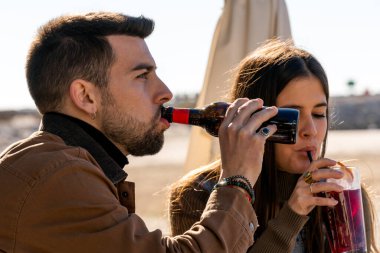 Young bearded male in outerwear drinking alcoholic beverage from glass bottle while woman sipping cold cocktail through straw on beach
