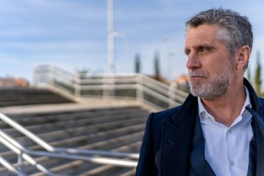 Confident bearded senior businessman in formal wear looking away while standing on stairs and thinking about project during walk