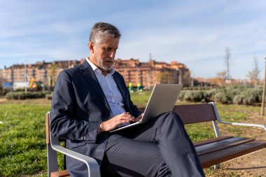 Focused mature businessman in formal suit working on project on laptop while sitting on bench on street