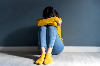 Full body of unrecognizable unhappy young lady with dark hair in casual clothes embracing knee and covering face while sitting on floor near wall