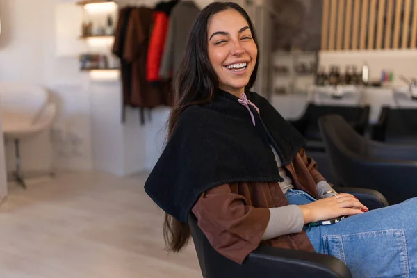Optimistic female customer laughing and looking at camera while sitting in armchair and waiting for master in modern salon