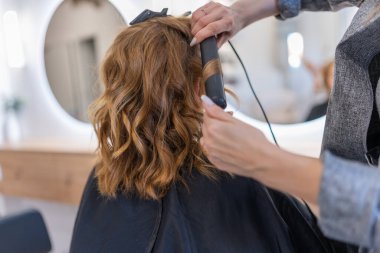 Unrecognizable hairdresser using professional tool to style hair of anonymous client in hairdressing chair against mirror