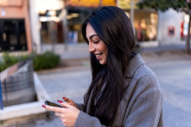 Optimistic young female with long dark hair wearing warm coat smiling and messaging on mobile phone while walking on street in city