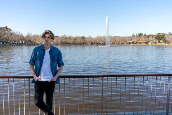Calm male in casual outfit with hands in pockets leaning on railing while standing against lake with fountain on sunny day