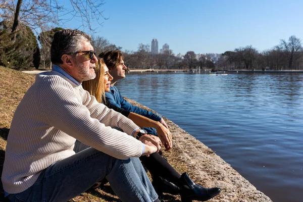 Side view full body of mature man and woman resting on river bank with son near leafless tree while enjoying peaceful weekend