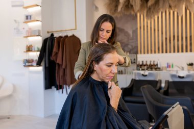 Side view of thoughtful mature female client with hand on chin using tablet while hairdresser combing hair in beauty salon