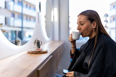 Side view of adult female client with long wet hair in black cape sitting in front of round mirror in modern beauty studio 