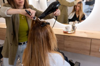 From above of crop anonymous female hairstylist in blazer drying hair of female customer in front of round mirror while working in modern salon