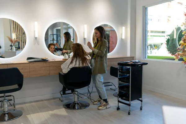 Full body of concentrated female hairdresser drying hair of client sitting on black chair in front of round mirror in light spacious beauty studio
