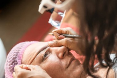 Anonim kadın kozmetik uzmanının, güzellik salonundaki müşteriye iplikle kaş şekli düzeltmesi yaptığı yüksek açı.