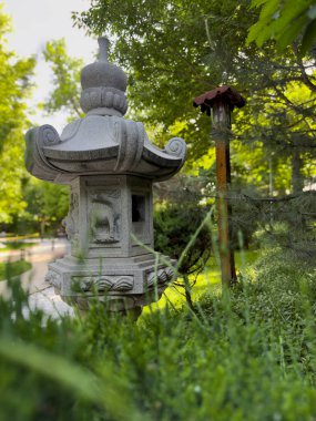 Geleneksel bir Japon taş feneri. Serin bir parktaki yemyeşil alanlar arasında kısmen saklı. Barışçıl atmosfer ve doğal ışık, doğanın ve geleneksel tasarımın uyumunu vurgular.