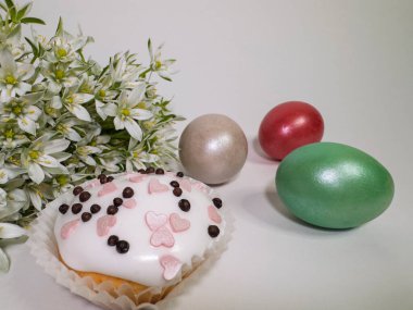 Beyaz kremalı Paskalya keki ve beyaz arka planda renkli yumurta ve bahar çiçekleri serpiştirilmiş.