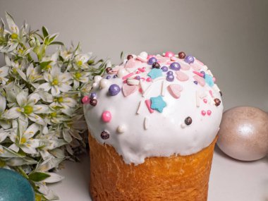 Paskalya pastası, şekerli krema, renkli serpiştirmeler, dekore edilmiş yumurtalar ve açık arkaplanda beyaz bahar çiçekleri. Geleneksel neşeli natürmort