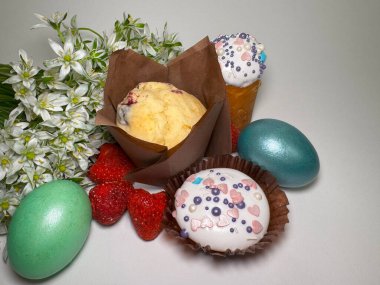 Paskalya temalı ev yapımı kekler ve kekler, beyaz krema ve renkli süslemelerle süslenmiş, etrafı boyanmış yumurta, çilek ve taze bahar çiçekleriyle çevrili.